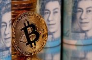 Moeda de Bitcoin dourada, com reflexo, pilhas de moedas e notas de libra esterlina desfocadas ao fundo.