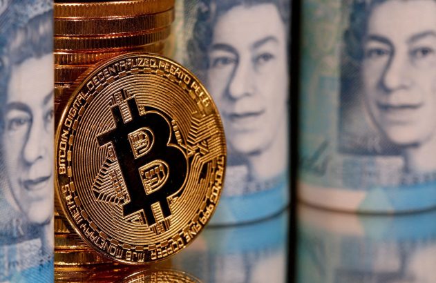 Moeda de Bitcoin dourada, com reflexo, pilhas de moedas e notas de libra esterlina desfocadas ao fundo.