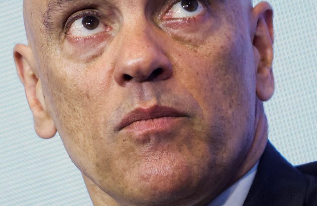 Homem careca de terno e gravata roxa. Ele olha atentamente para cima, com fundo colorido e desfocado.