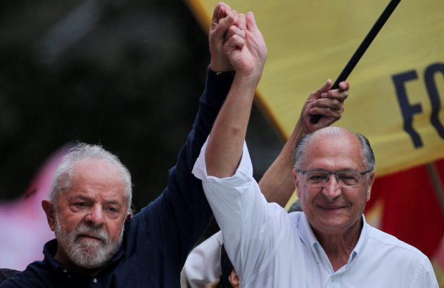 Lula e Alckmin, com mãos unidas e erguidas. Alckmin sorri e Lula tem expressão séria. Fundo desfocado.