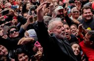 Lula, de barba e cabelo branco, levanta o braço, cercado por uma multidão sorridente com celulares em punho.
