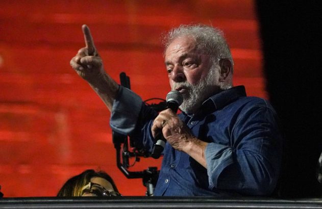 Lula, com barba grisalha e camisa azul, discursa ao microfone com o dedo indicador para cima em fundo vermelho.