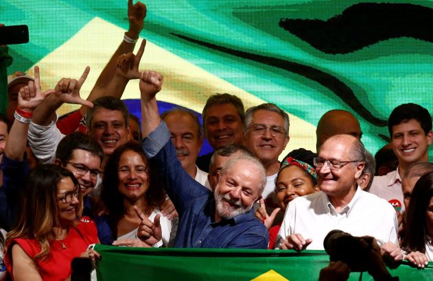 Lula sorri e ergue o punho em meio a apoiadores, com bandeiras do Brasil ao fundo e à frente.