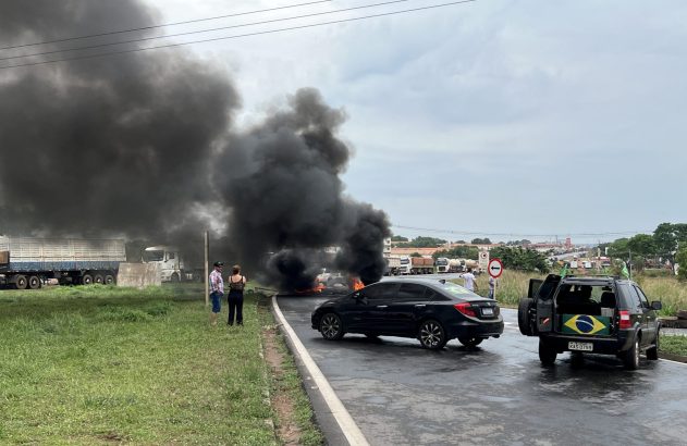 Fogo e fumaça preta intensa bloqueiam rodovia. Carros e pessoas observam. SUV com bandeira do Brasil.