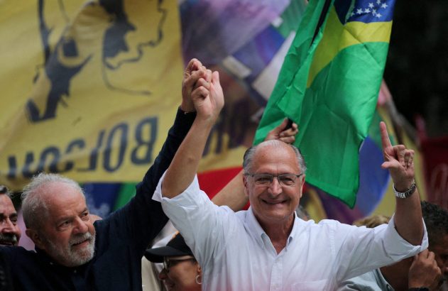 Dois homens sorrindo e com braços erguidos, um segurando a mão do outro. Ao fundo, bandeiras.