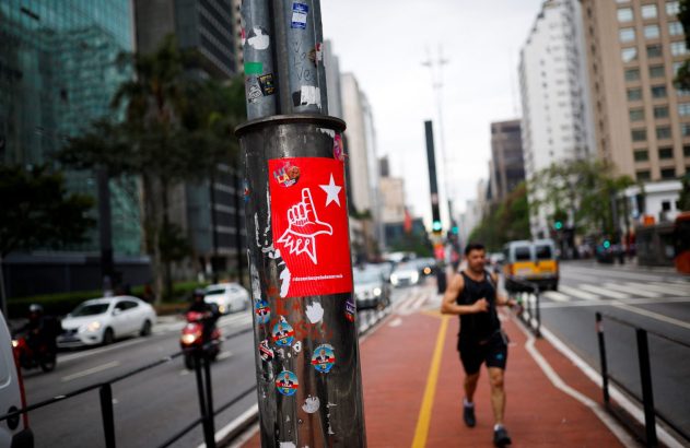 Poste com adesivos, um vermelho com mão e estrela. Homem corre em calçada vermelha de avenida urbana com tráfego e prédios.