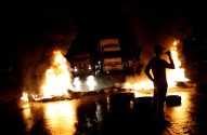 Bloqueio noturno: Fogo na rua molhada, caminhões parados e pessoas em silhueta. Reflexos laranja.