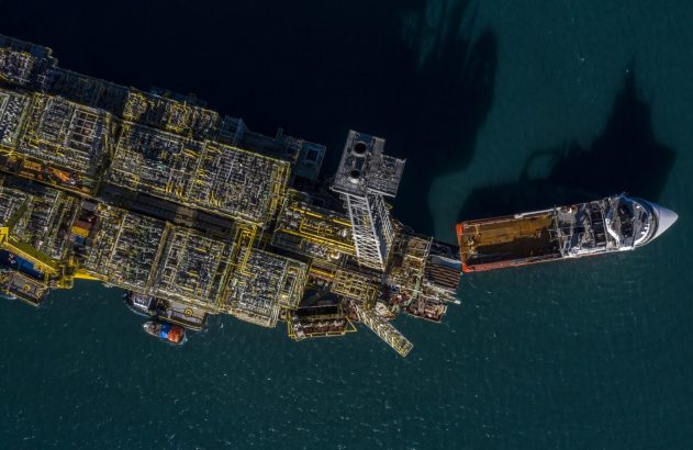 Vista aérea de uma grande plataforma offshore flutuante com tubulações amarelas, e um navio de suporte atracado em águas oceânicas.