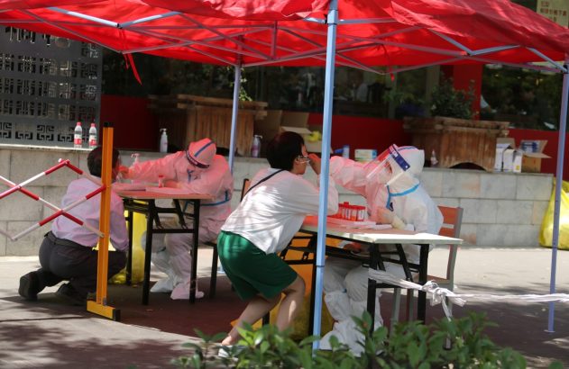 Pessoas em trajes de proteção brancos realizam testes em civis sob uma tenda vermelha.