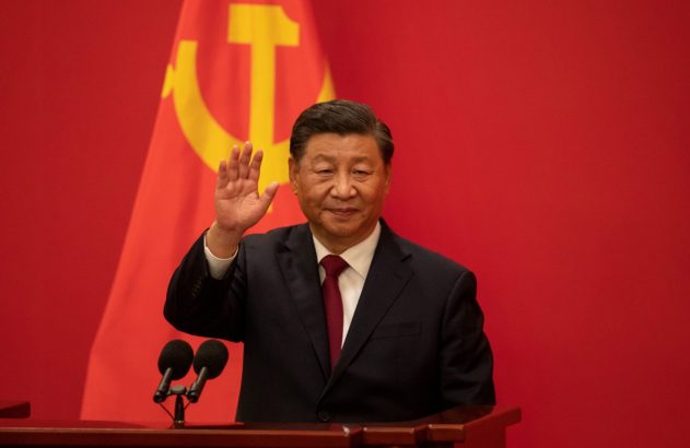 Xi Jinping, líder chinês, acena em pódio com emblema do PCC em fundo vermelho.