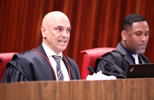 Dois magistrados com togas. Um fala, o outro usa um laptop em audiência.