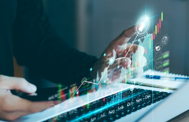 A imagem mostra as mãos de uma pessoa, que está sentada em frente a um notebook enquanto analisa ações ordinárias e preferenciais. Um gráfico de candlestick foi sobreposto digitalmente na imagem. (Foto: Shutterstock / Thapana_Studio)
