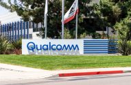 Sinal da Qualcomm em branco e azul em frente ao prédio da empresa. Gramado verde e bandeira da Califórnia visíveis.