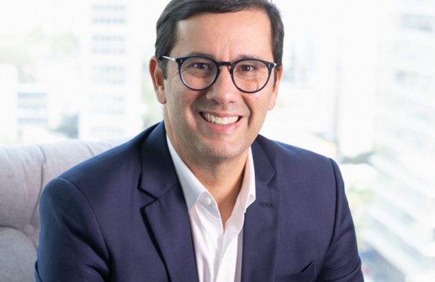 Homem sorridente de óculos e blazer azul, em ambiente profissional.