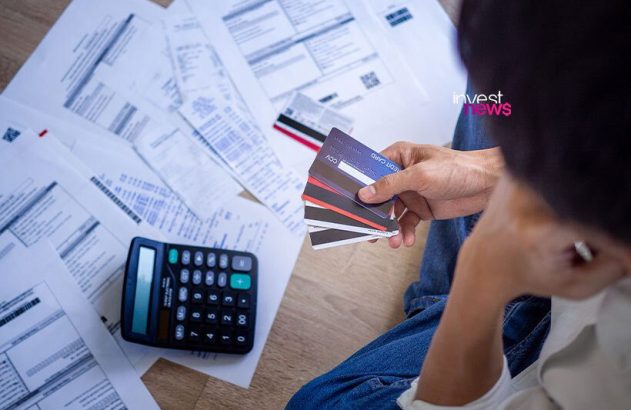 Mão segura vários cartões de crédito junto a contas e calculadora no chão. Cena de dívidas e estresse financeiro.