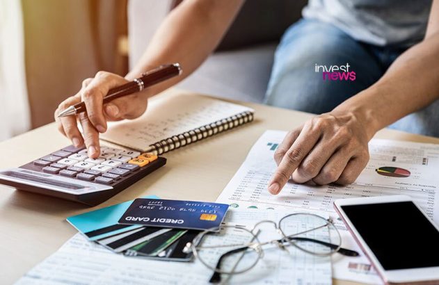 Mãos usando calculadora e analisando planilha de despesas. Mesa com óculos, cartões de crédito e celular em contexto financeiro.