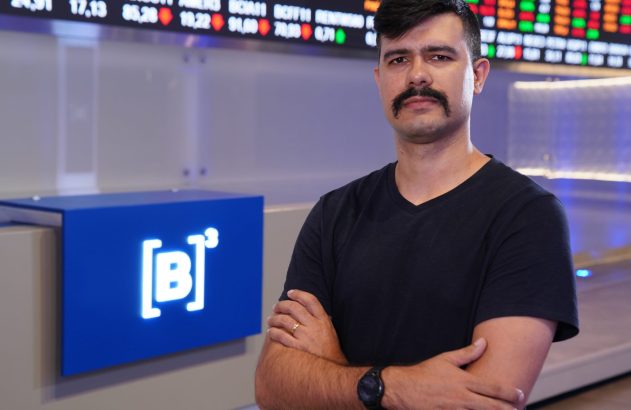 Homem de bigode e braços cruzados no ambiente da B3, com painel de dados financeiros ao fundo.