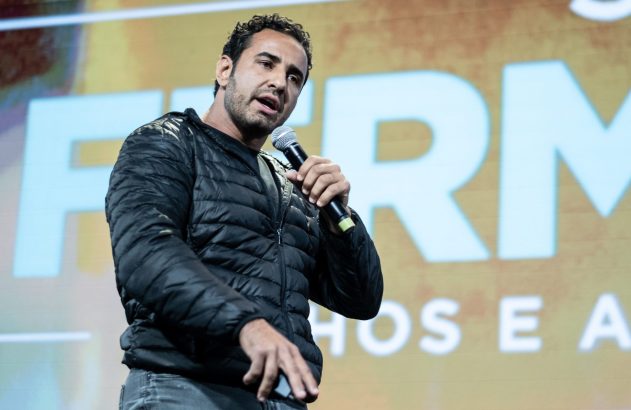 Homem de jaqueta preta fala ao microfone em palco, com letreiro ao fundo.