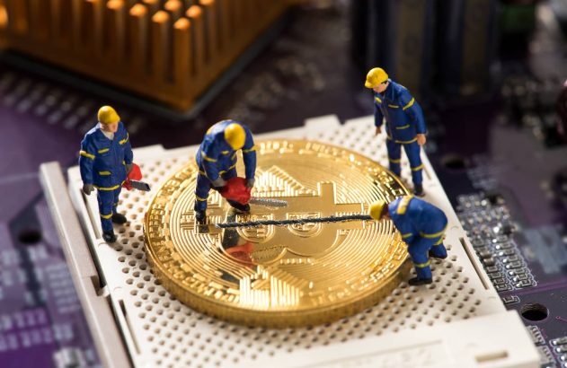 A imagem mostra uma moeda que simboliza o bitcoin em cima do que parece ser uma placa de computador. Há quatro bonecos, vestidos com macacões azuis e chapéus amarelos, serrando a moeda ao meio, ilustrando o processo de halving. (Imagem: Shutterstock / PowerUp)