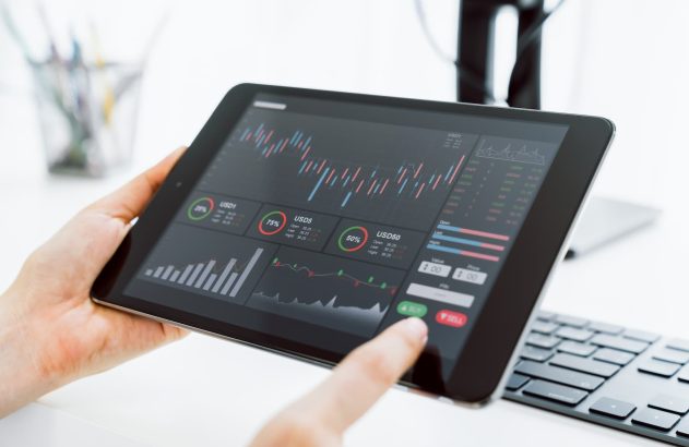 A imagem mostra as mãos de uma pessoa branca segurando um tablet preto. Na tela do site, vemos uma home broker com diversos gráficos, indicando que a pessoa possa estar analisando operações em intraday. (Foto: Shutterstock / sitthiphong)