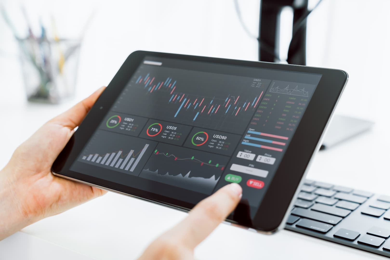 A imagem mostra as mãos de uma pessoa branca segurando um tablet preto. Na tela do site, vemos uma home broker com diversos gráficos, indicando que a pessoa possa estar analisando operações em intraday. (Foto: Shutterstock / sitthiphong)