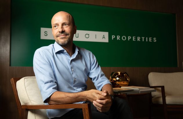 Homem careca de barba e camisa azul sorri sentado. Fundo: logo 'SEQUOIA PROPERTIES' em parede verde.