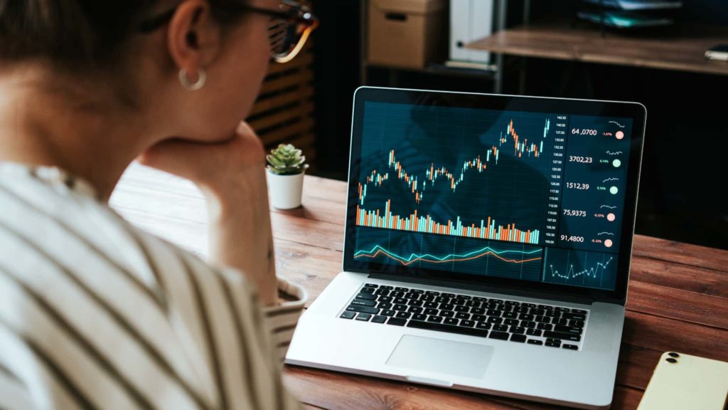 Mulher branca, usando uma blusa listrada e óculos, observa gráficos de cotação representando operações swap na tela de um notebook prateado. (Foto: Shutterstock / Alex from the Rock)