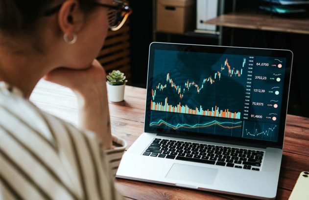 Mulher branca, usando uma blusa listrada e óculos, observa gráficos de cotação representando operações swap na tela de um notebook prateado. (Foto: Shutterstock / Alex from the Rock)