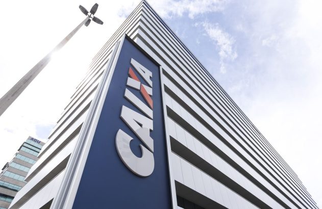 Edifício alto com logo da CAIXA, visto de baixo. Poste de luz à esquerda e céu claro com nuvens.