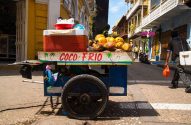 Carrinho de coco frio cheio de cocos e gelo em rua urbana. Uma pessoa caminha ao lado de edifícios coloridos.