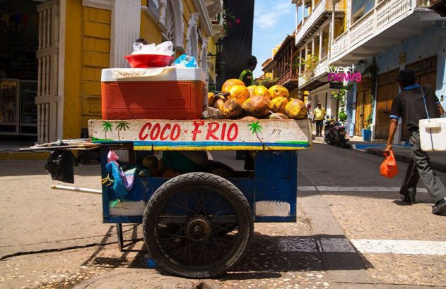 Carrinho de coco frio cheio de cocos e gelo em rua urbana. Uma pessoa caminha ao lado de edifícios coloridos.