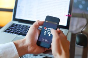 Mãos segurando smartphone com app de compras: carrinho com 2 itens e botão "BUY NOW". Laptop e mouse ao fundo.