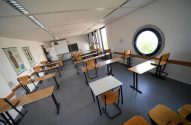 Sala de aula com carteiras e cadeiras. Uma pessoa, quadros anatômicos na parede e uma janela redonda.
