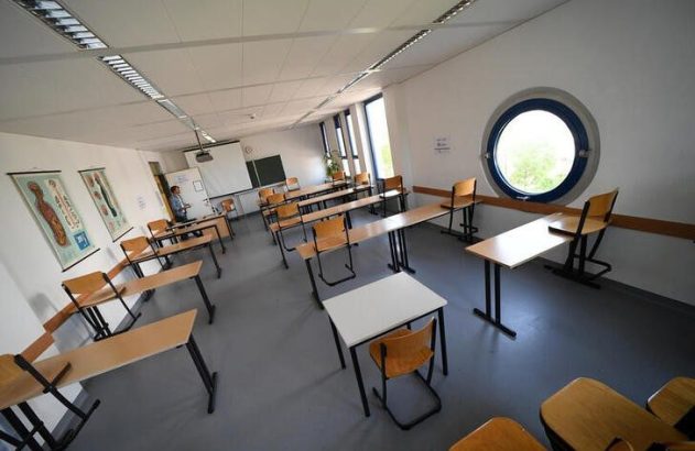 Sala de aula com carteiras e cadeiras. Uma pessoa, quadros anatômicos na parede e uma janela redonda.