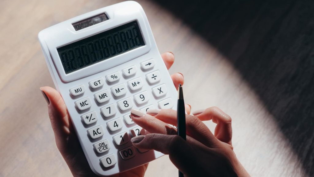 A imagem mostra um par de mãos brancas, com as unhas pintadas, fazendo contas em uma calculadora branca. (Foto: Pexels / Kindel Media)