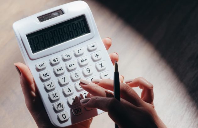 A imagem mostra um par de mãos brancas, com as unhas pintadas, fazendo contas em uma calculadora branca. (Foto: Pexels / Kindel Media)