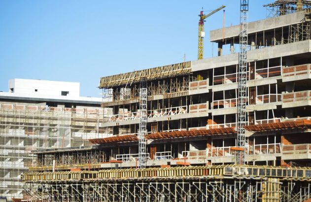 Vários edifícios em construção com estruturas de concreto expostas, andaimes e guindastes sob céu azul.