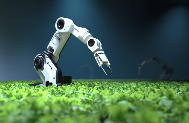 Braço robótico branco com pinças sobre uma plantação verde. Outro robô desfocado ao fundo.