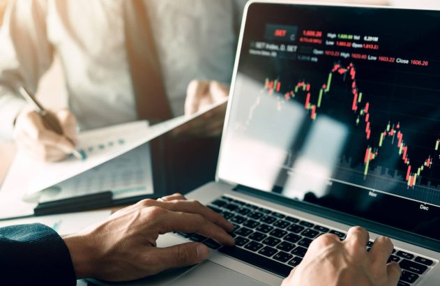 A imagem mostra, em primeiro plano, as mãos de uma pessoa digitando em um notebook prateado, enquanto a tela exibe um gráfico de candlestick. No fundo da imagem, outra pessoa analisa dados impressos em folhas de papel. (Foto: Shutterstock / wutzkohphoto)