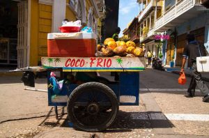 Carrinho de coco frio cheio de cocos e gelo em rua urbana. Uma pessoa caminha ao lado de edifícios coloridos.