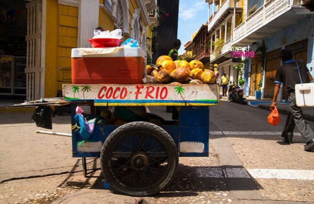 Carrinho de coco frio cheio de cocos e gelo em rua urbana. Uma pessoa caminha ao lado de edifícios coloridos.