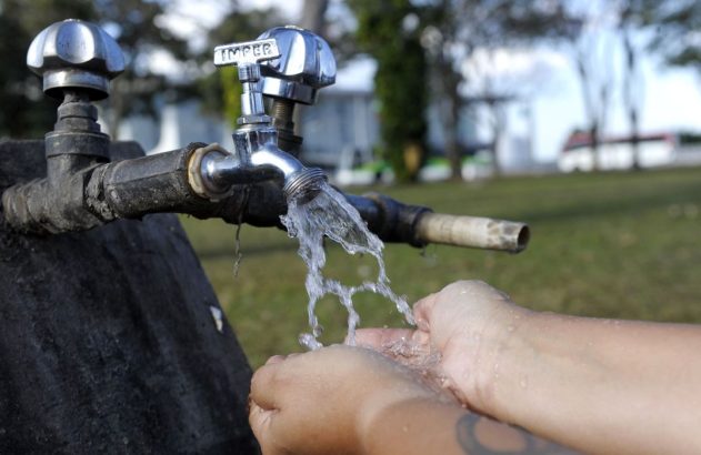 Mãos em concha recebem água de torneira externa com marca 'IMPER'. Água escorre, fundo verde e desfocado.