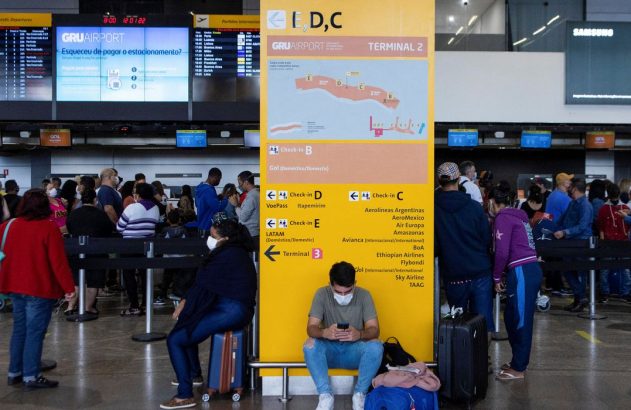 Muitas pessoas com máscaras esperam em terminal de aeroporto, com telas de informação e um grande sinal de check-in.