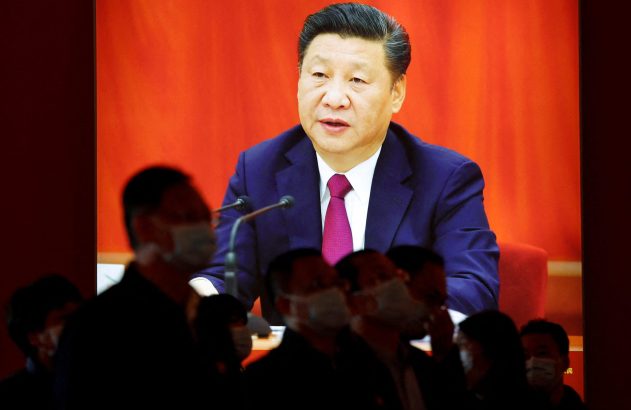 Xi Jinping discursando em telão com fundo vermelho. Pessoas em silhueta, algumas de máscara, assistem.