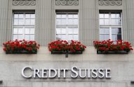 Letreiro 'Credit Suisse' em prédio cinza com janelas e floreiras de flores vermelhas.
