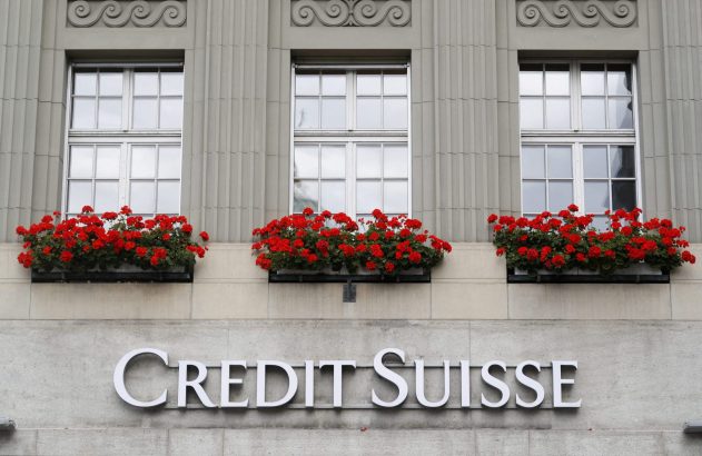Letreiro 'Credit Suisse' em prédio cinza com janelas e floreiras de flores vermelhas.