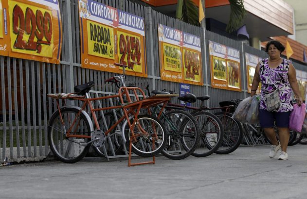 Mulher com sacolas caminha. Bicicletas estacionadas e cartazes de preços em cerca de loja.