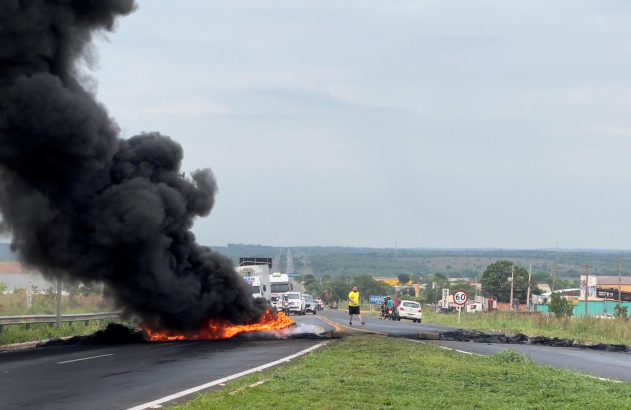 Rodovia bloqueada por pneus queimando, com intensa fumaça preta. Pessoas e veículos detidos.