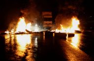 Caminhão à noite entre pneus em chamas bloqueando estrada molhada, com reflexos de fogo no chão.
