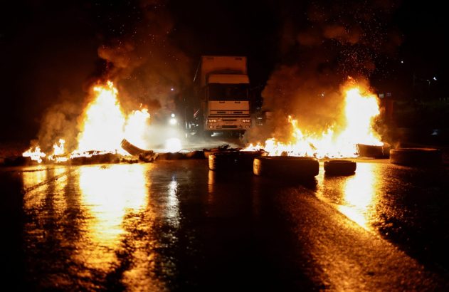 Caminhão à noite entre pneus em chamas bloqueando estrada molhada, com reflexos de fogo no chão.
